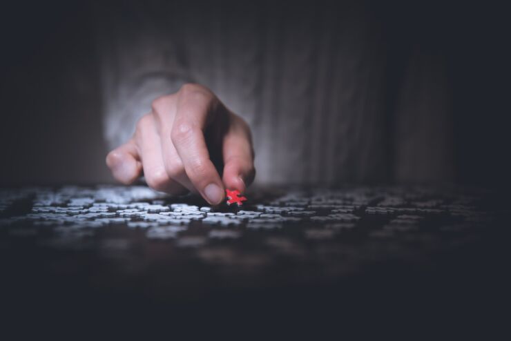 A close up on a hand that holds a piece of a puzzle. Under the hand there is a lot of other pieces of a puzzle.