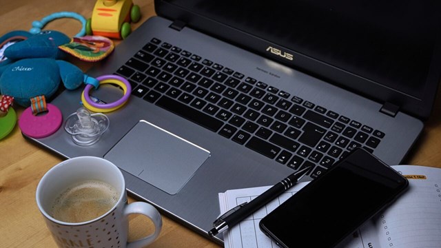En närbild på en dator. Runt datorn ligger olika saker; till vänster två leksaker, rakt framför en kopp med kaffe och till höger ligger en anteckningsbok och en mobil.