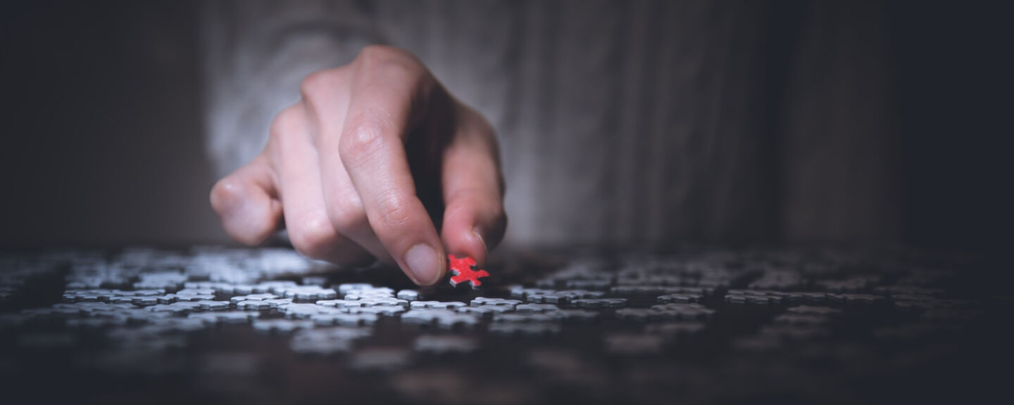 Närbild på en hand som håller i en röd pusselbit vid ett bord. Under handen ligger flera vita pusselbitar.