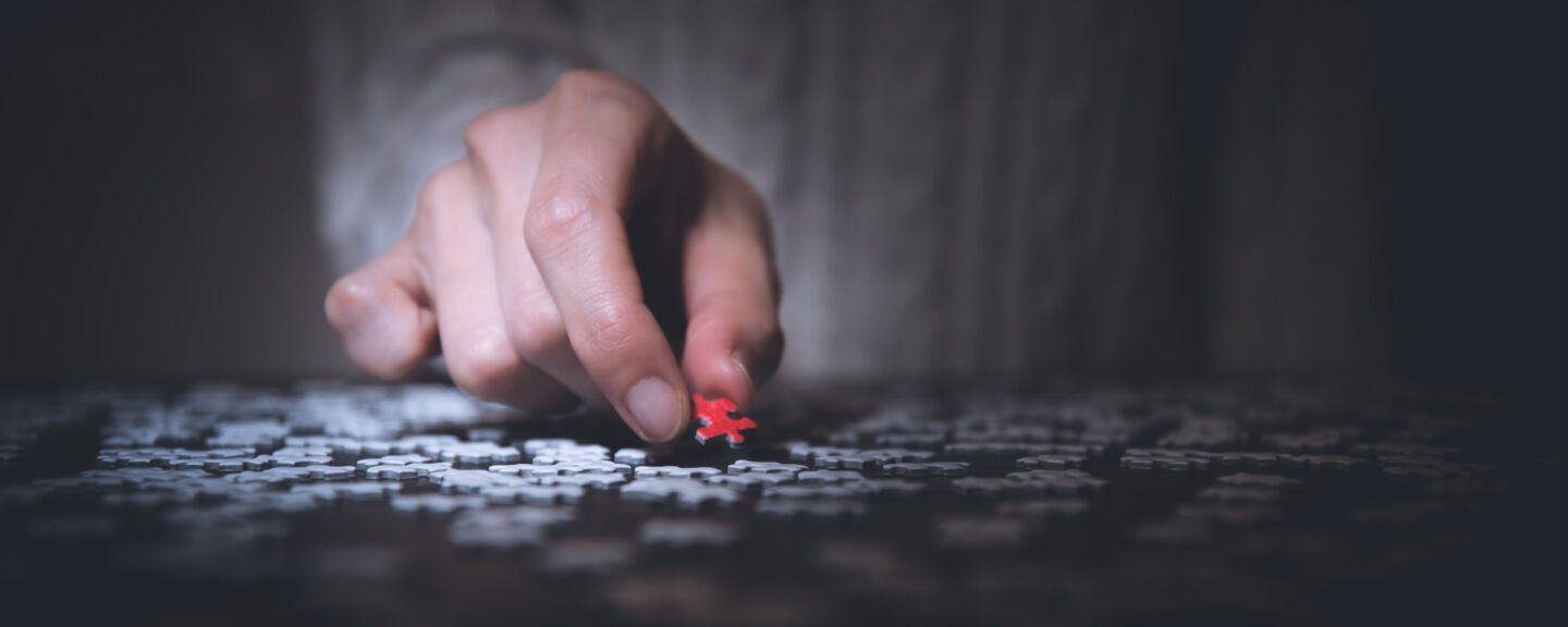 Close up of a hand holding a red piece of puzzle. Under the hand there are a lot more pieces, but they are white.