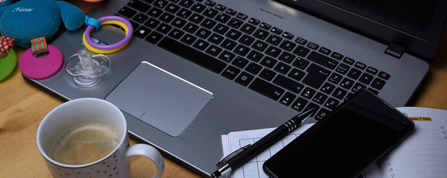 Close up of a laptop. On the left side of the laptop there are two toys, in front of the keyboard there is a cup of coffee and on the left there is a note pad and a mobile phone.