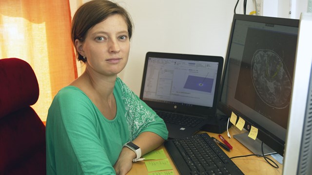 Teresa Nordin sitter vid sitt skrivbord med tre skärmar och ett tangentbord framför sig. Hon har en grön tröja och på den mittersta skärmen syns en bild på en hjärna.