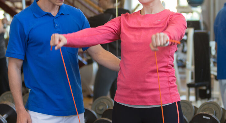 A physiotherapist in a blue shirt is standing to the left of a woman in a pink shirt doing an exercise. You can not see their faces.