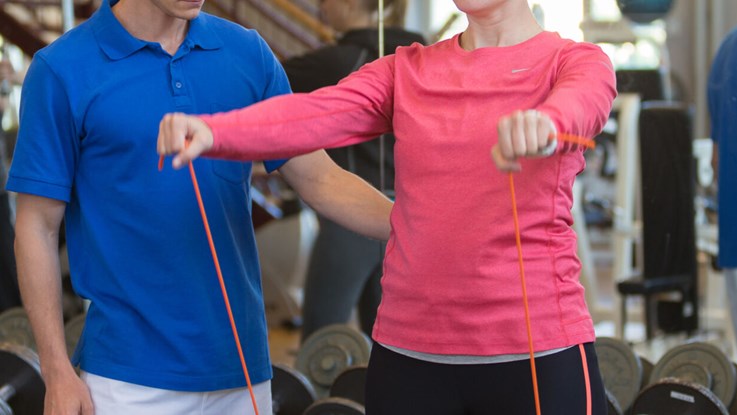 A physiotherapist in a blue shirt is standing to the left of a woman in a pink shirt doing an exercise. You can not see their faces.