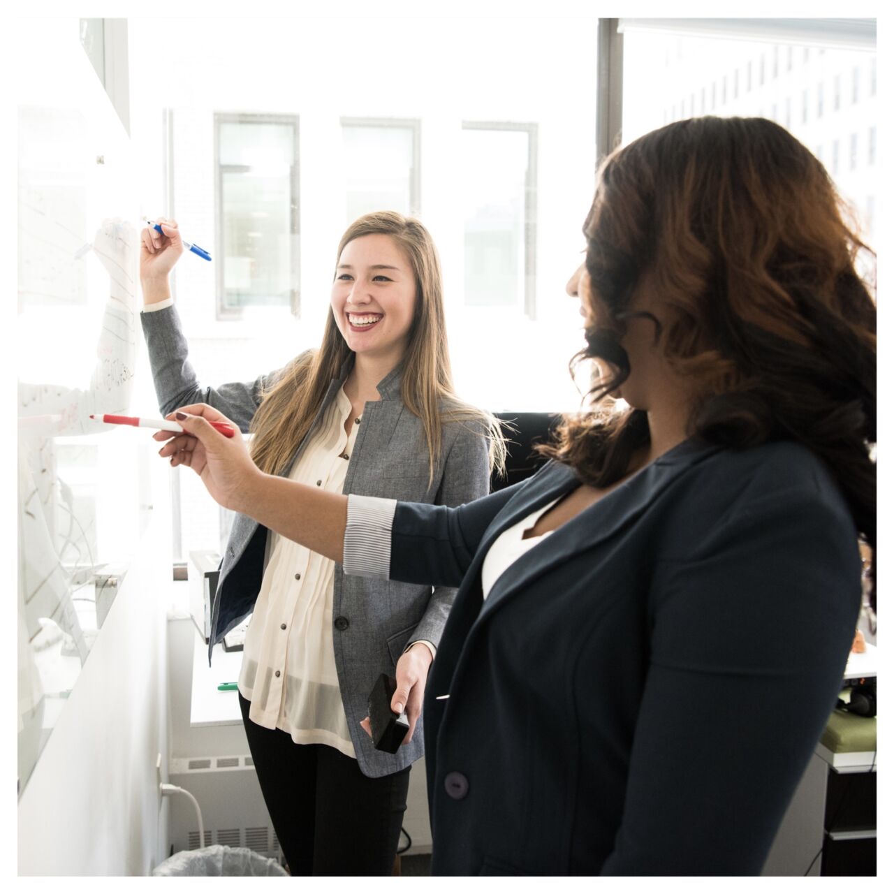  Two laughing women standing and writing on whiteboard.
