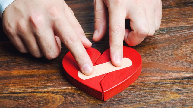 A pair of hands are putting a plaster on a red, flat wooden heart.