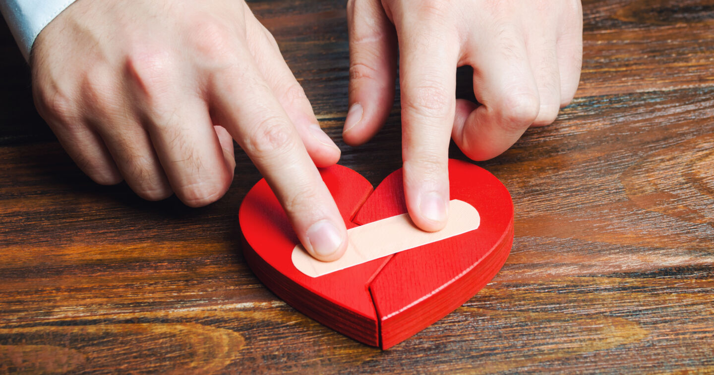 Ett rött, platt trähjärta ligger på ett bord. Ett par händer sätter ett plåster på det.
