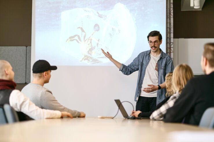 Studenter vid ett bord. En person stor vid bordet och presenterar något på storbild.