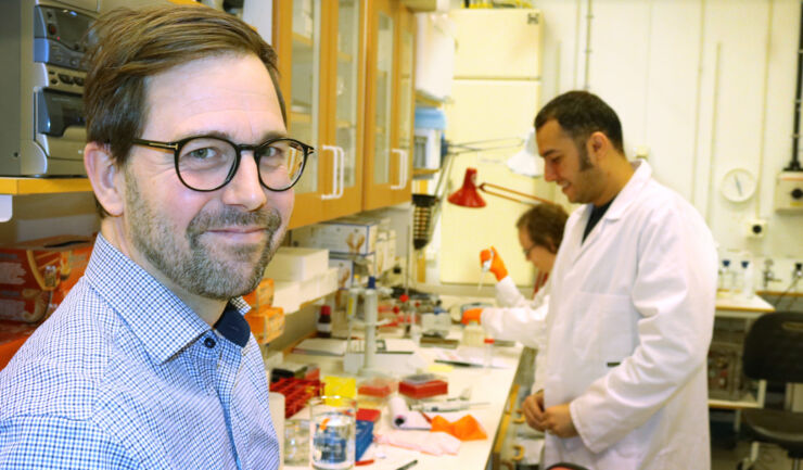 Man in lab, looking into the camera, in the background a female and a male PhD student.