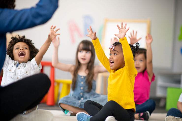 Barn i förskolan sitter på golvet framför förskolläraren och räcker ivrigt upp händerna.