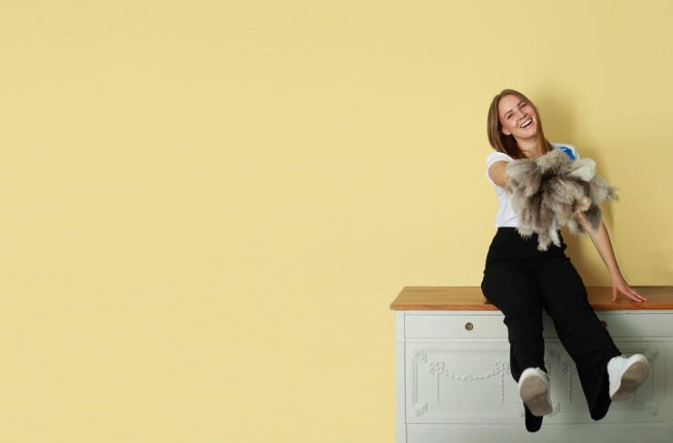 Clara Johansson sitter på en grön byrå mot en gul vägg och ler. I handen har hon en dammvippa.