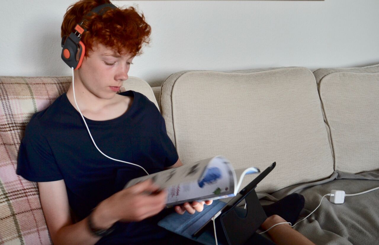 Pojke sitter i soffan med hörlurar och surfplatta med en tidning i handen. 