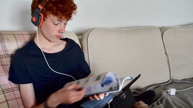 Pojke sitter i soffan med hörlurar och surfplatta med en tidning i handen. 