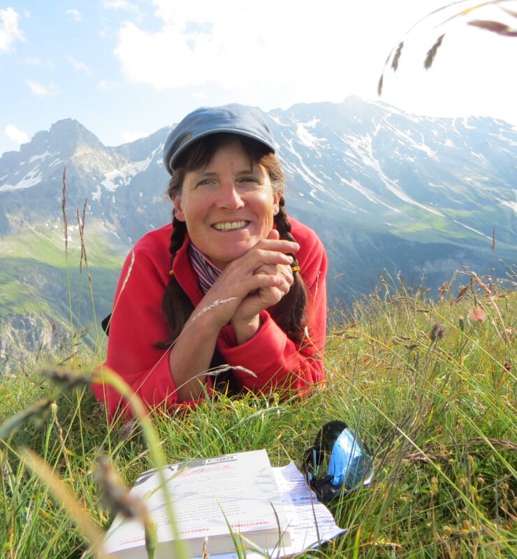 Alice Henderson laying in the green grass with a book. Mountins are towering behind her.