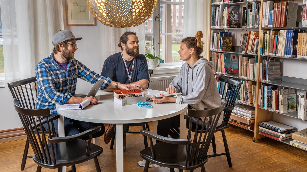 Three students at a round table in front of a book shelf.