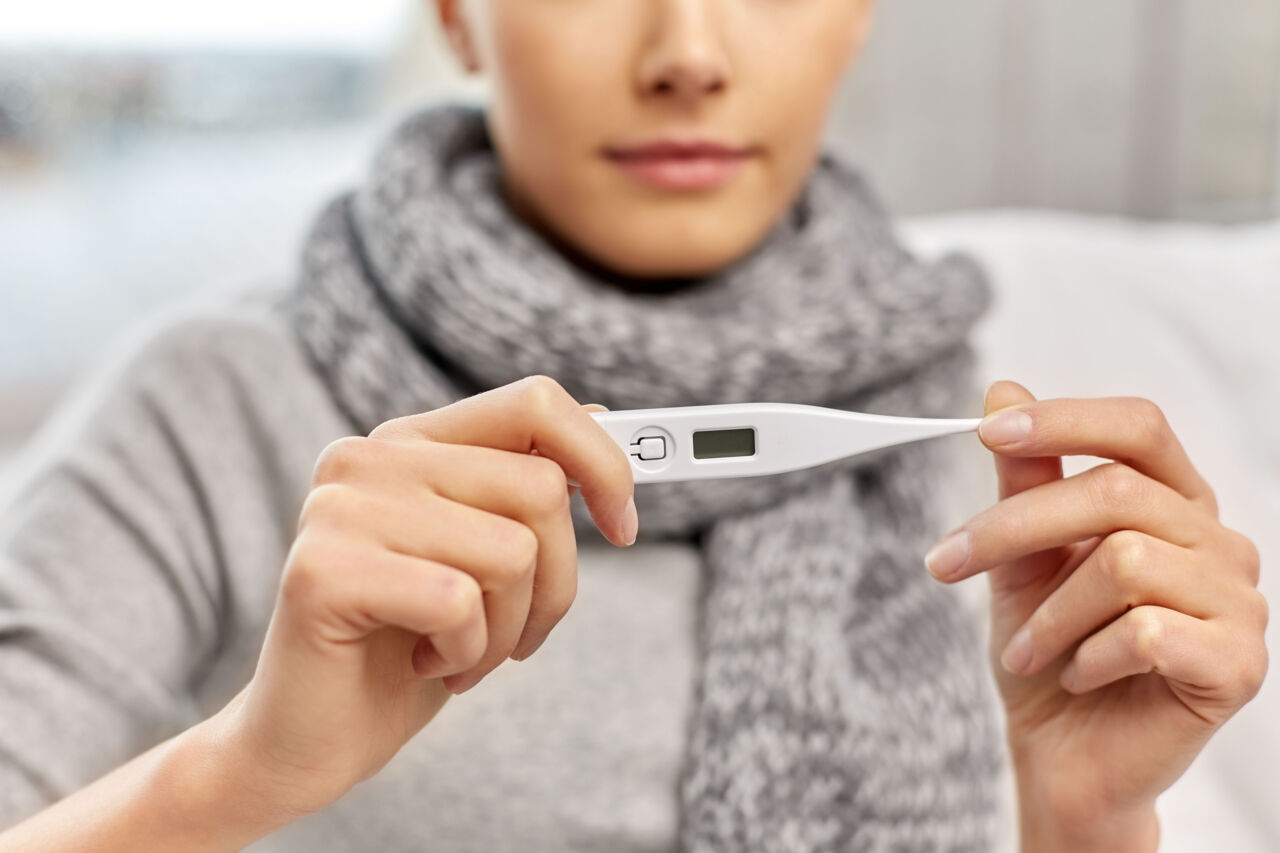 En person i grå tröja och grå halsduk håller i en febertermometer.