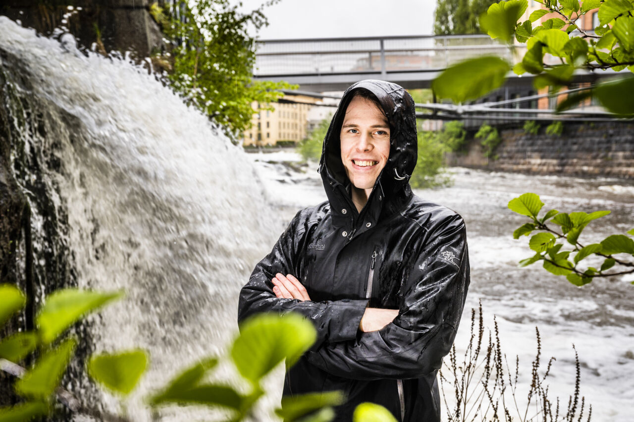 En man iklädd svart regnrock med uppfälld luva står framför ett vattenfall