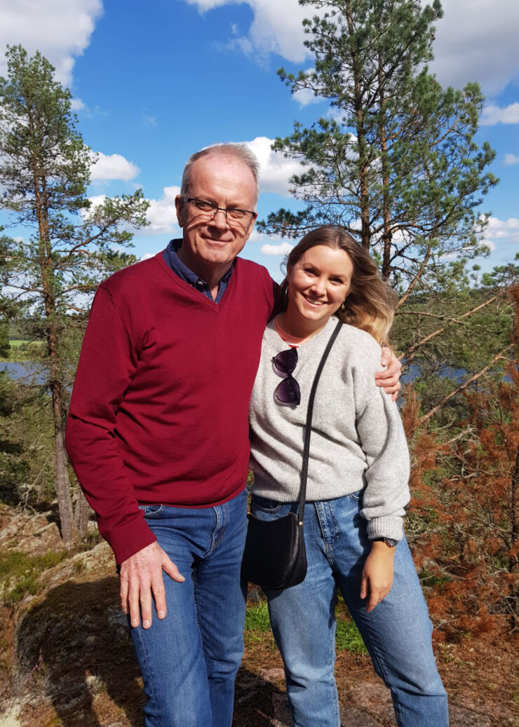 En man och en yngre kvinna står och håller om varandra utomhus. Mannen har röd tröja. Det är träd bakom dem. De ser glada ut.