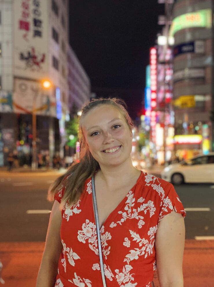 Young woman, wearing ar red and white dress, standing in front of buildings with neon lights