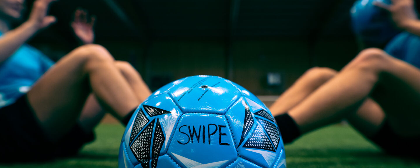 Close up of a blue football. In the background four persons are doing sit ups.