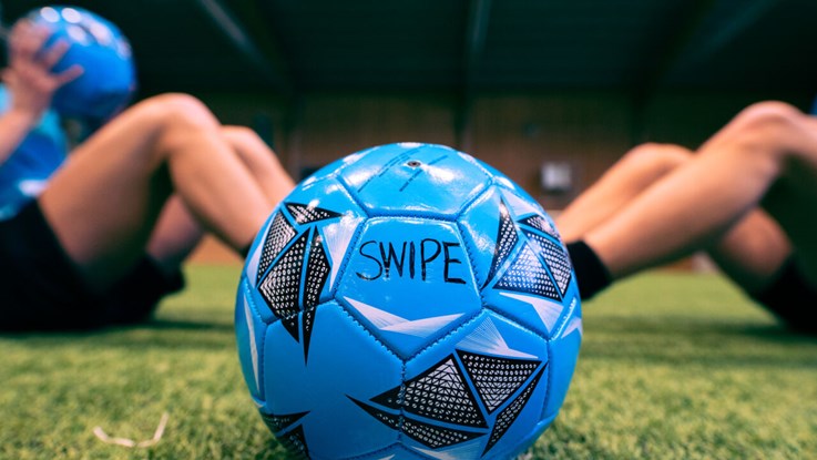 Close up of a blue football. In the background four persons are doing sit ups.