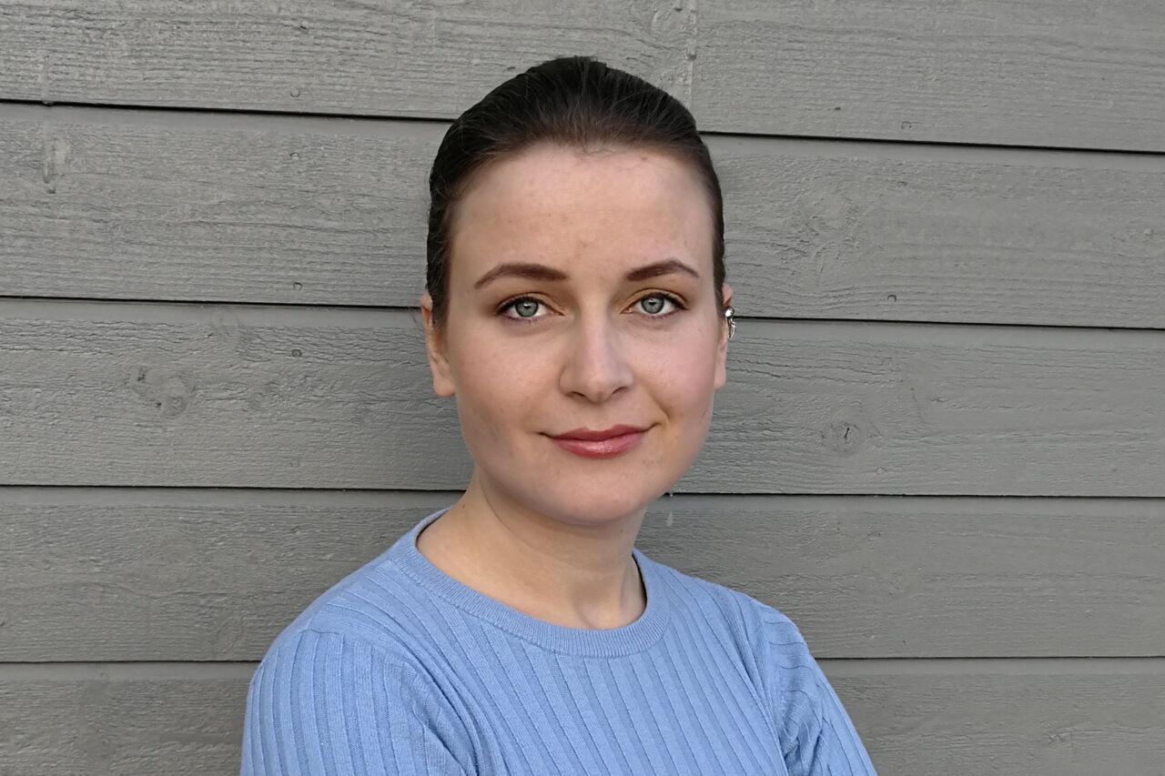 Porträttbild av mörkhårig ung kvinna i ljusblå tröja mot grå vägg