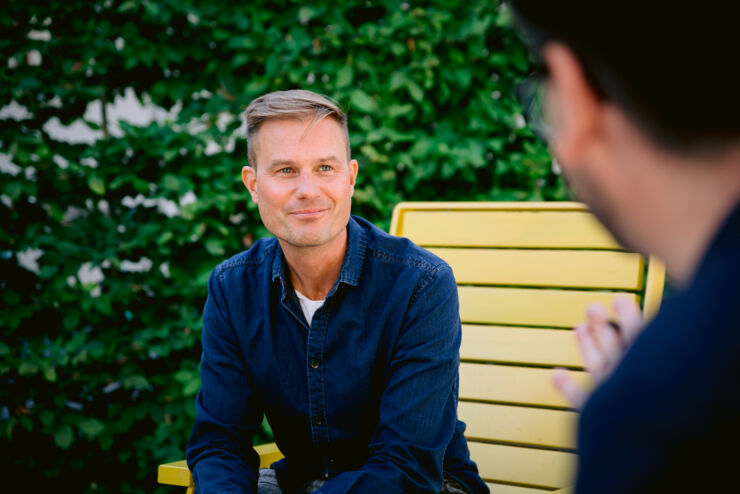 Man sits outside. Back of another man's head at the edge of the picture.