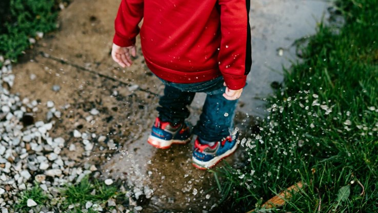 Närbild på ett barn som hoppar i en vattenpöl. Barnet har en röd tröja och jeans på sig.