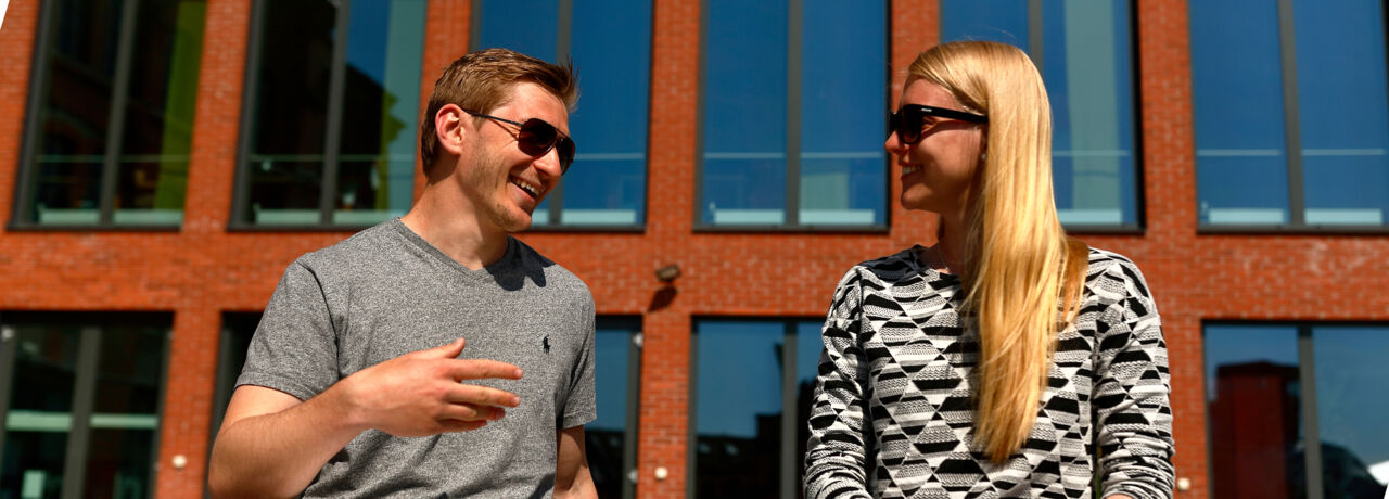 Närbild av två studenter med solglasögon framför en tegelbyggnad med stora fönster