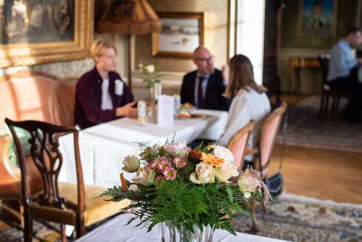 Foto på en blombukett på ett pelarbord med tre suddiga personer i bakgrunden.