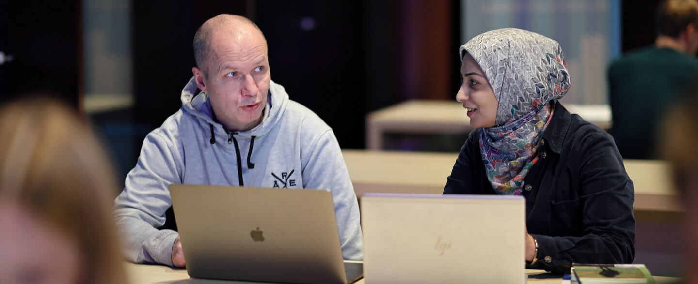 En man och en kvinna i slöja sitter och pratar vid ett bord inne på biblioteket. De har böcker och datorer på bordet. 