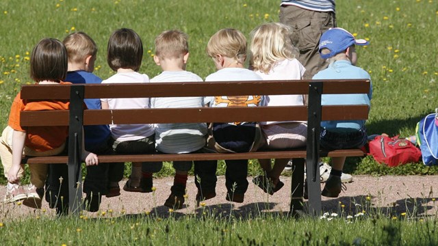 Förskolebarn sitter på rad på en bänk ute i solen.