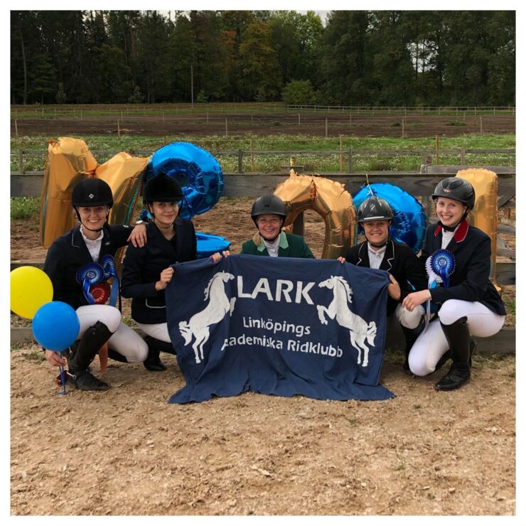 Four girls in riding clothes sitting on the ground with blue and yellow ballongs and a LARK-sign.