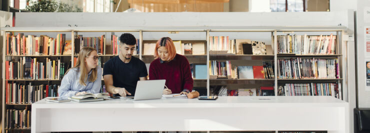 Tre studenter sitter i ett bibliotek och studerar.