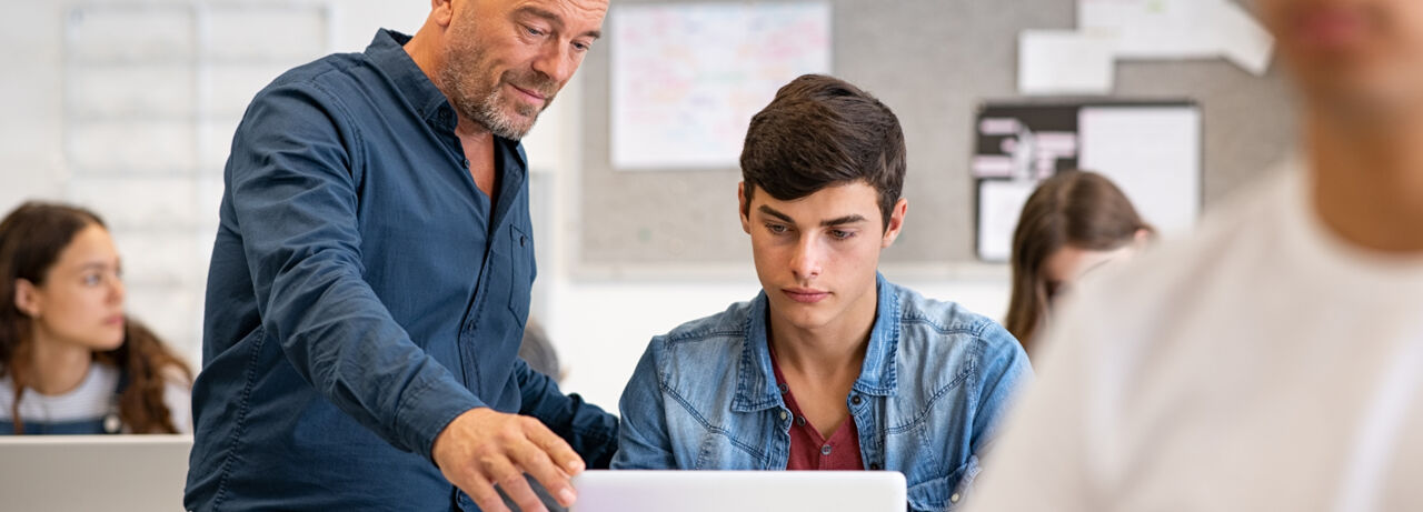 En lärare står bredvid och hjälper en elev som sitter vid ett skrivbord med en bärbar dator på. 