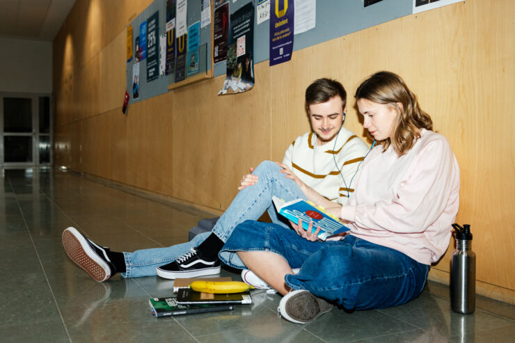 Två studenter sitter på golvet i en korridor och läser i en bok och lyssnar på musik