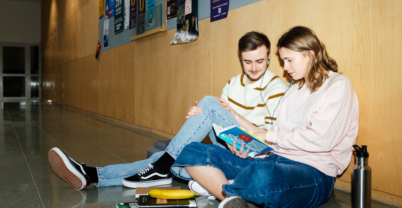 Två studenter sitter på golvet i en korridor och läser i en bok och lyssnar på musik