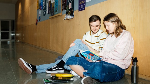 Två studenter sitter på golvet i en korridor och läser i en bok och lyssnar på musik