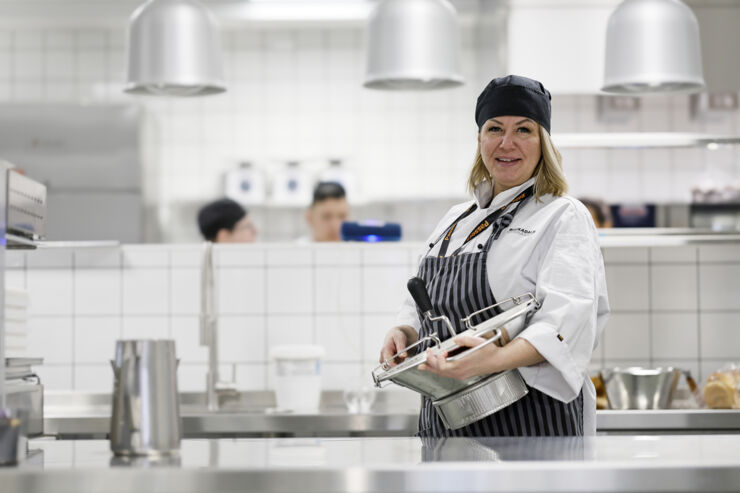 Kvinna i kockmössa står i ett restaurangkök och tittar in i kameran