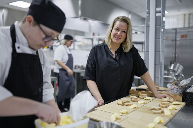 Kvinna i kockmössa står i ett restaurangkök och tittar in i kameran. I förgrunden står en kockkollega.