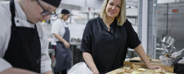 Kvinna i kockmössa står i ett restaurangkök och tittar in i kameran. I förgrunden står en kockkollega.