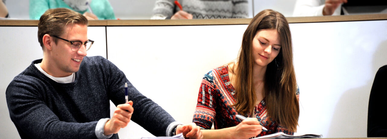 Närbild av ung man med glasögon och ung kvinna med långt brunt hår i en föreläsningssal.