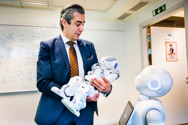 The minister is holding a little white robot while a larger white service robot is standing on the floor looking up at him.