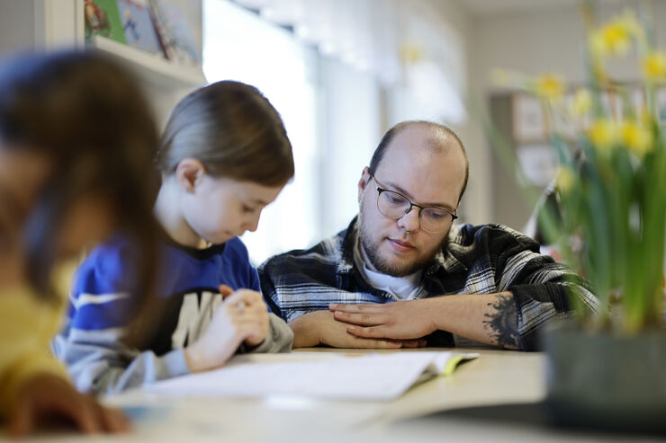 A student sits at his school desk and is helped by a teacher squatting next to him.