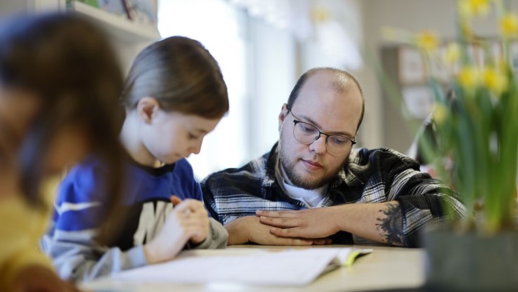 A student sits at his school desk and is helped by a teacher squatting next to him.