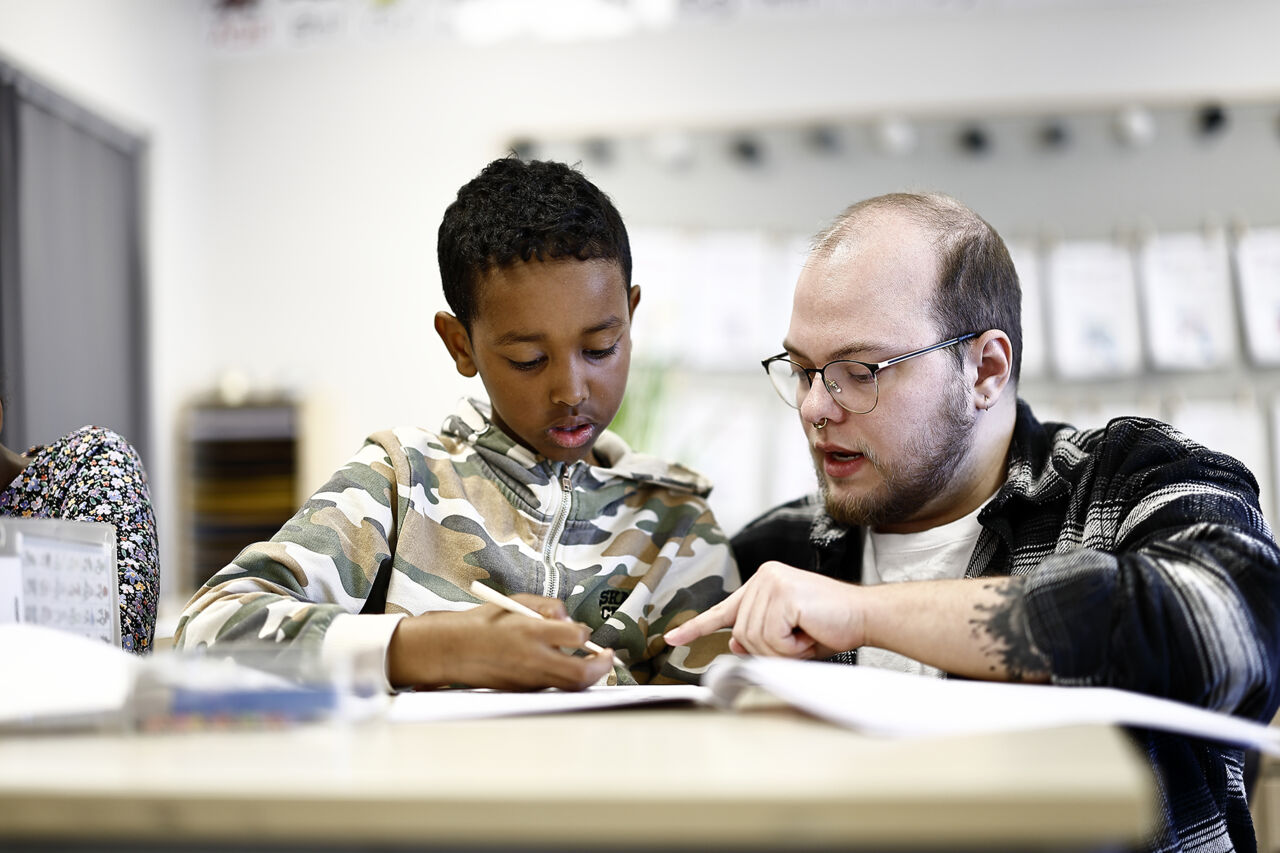 En lågstadieelev får hjälp av sin lärare som sitter på huk bredvid skrivbordet. De tittar ner och pekar i en bok.