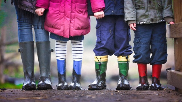 Fyra par ben på barn som har gummistövlar i olika färger. 