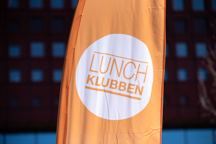 En beachflagga med Lunchklubbens logotyp på orange bakgrund. 
