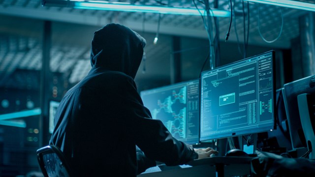 Person with hoodie sitting behind computer in dark room.