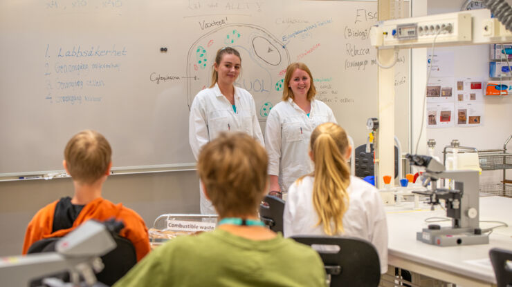 Två studenter står framför en grupp ungdomar i ett laboratorium.